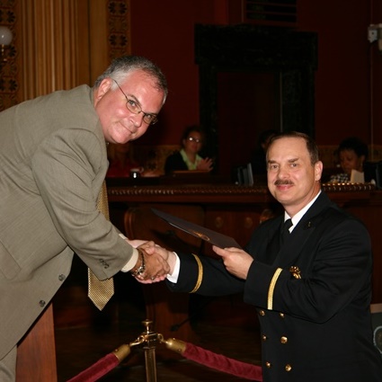 Lieutenant Legg in uniform being handed the Columbus City Council Honor and Recognition Award by Council President Mike Mentel wearing a tan suite and tie at the September 2005 Council meeting. 