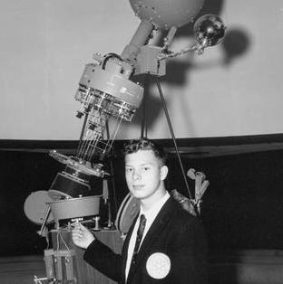 Charles Legg, wearing a suit and tie is standing at the base of the COSI Planetarium projector when he was 15 years old. 