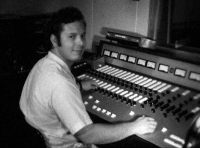 Charles is seated at a W O S U radio station control console in 1970. He is adjusting the knobs and slider controls on the control board as he is looking back over his shoulder. 