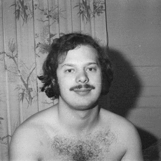 A young shirtless Charles sits in 1970 with long thick hair, a trimmed mustache, and hairy chest ready to take on the world.