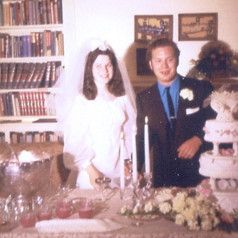Charles and Carol at their wedding reception in July of 1971. Carol is wearing a white wedding dress while Charles has a black suit and tie and a blue shirt. Also seen is the wedding cake and punch bowl.