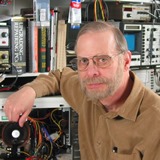 Charles Legg sitting at his electronics workbench with a computer in the background.